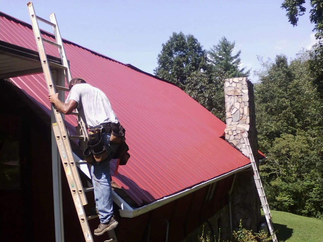 Roofer on a ladder beside a red metal roof and stone chimney — Soffit & Fascia Repair by Roofing Brothers in Berkeley Heights